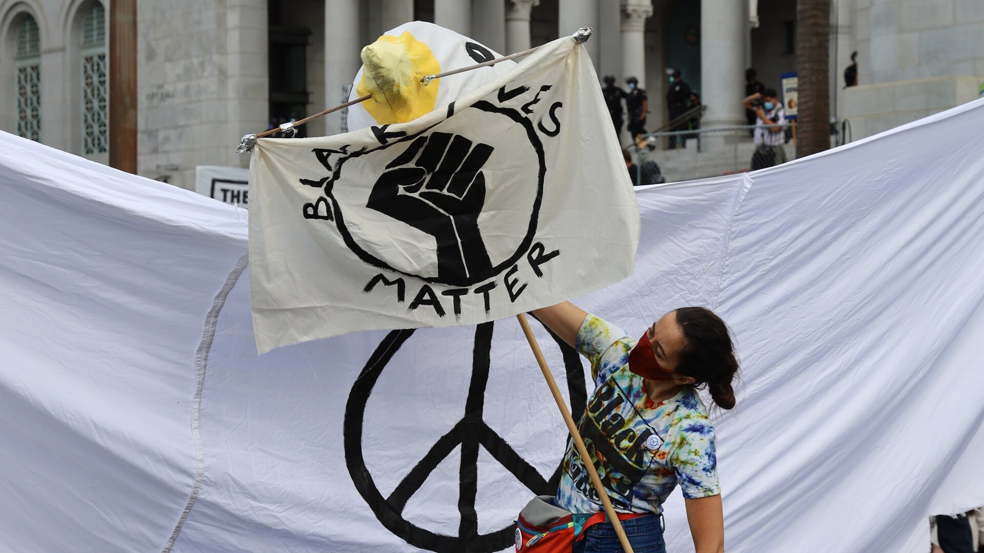 Foto van een vrouw op een demonstratie met een Black Lives Matter-bord. Achter haar is een spandoek met het peace-teken.